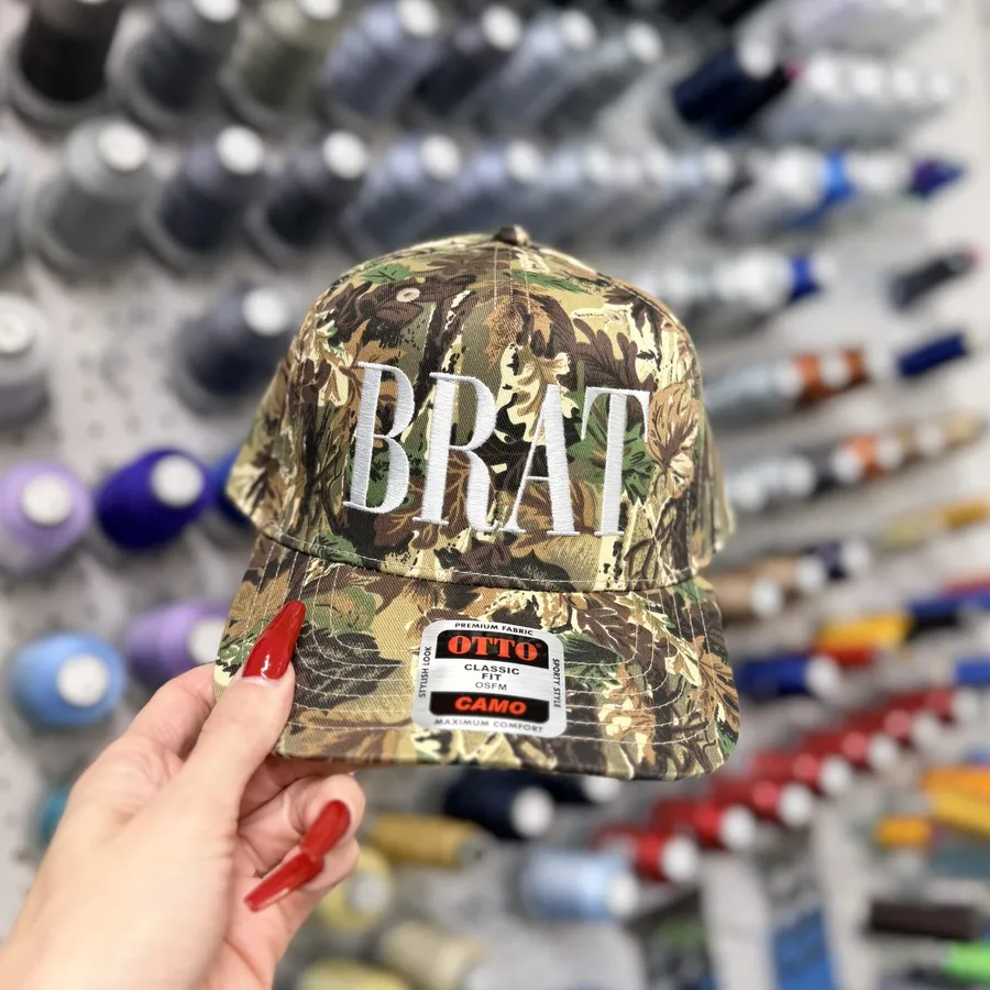 Embroidered camo cap held in front of thread racks at the Edge Embroidery shop